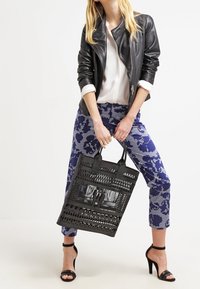 Svart perforerad tote-väska med utskärningsdesign, platta handtag. Bärs med mönstrade blå byxor och en svart läderjacka.