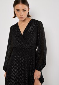 Kobieta z krótkimi włosami, ubrana w czarną sukienkę z długim rękawem w serek, pokrytą teksturowanym wzorem w zygzak, patrzy lekko w bok.