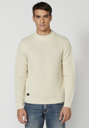 Jersey de punto crema con un patrón texturizado, cuello redondo ribeteado y puños. Combinado con jeans de mezclilla azules. Etiqueta marrón sutil en el dobladillo.