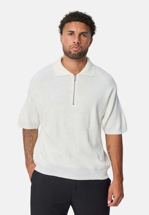 Homme aux cheveux bouclés se tient face à nous, portant un pull blanc à manches courtes en tricot avec une fermeture éclair devant et un pantalon noir.