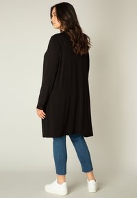 Zwarte langemouwen top met een relaxte fit en een langere achterkant, gecombineerd met blauwe cropped jeans en witte sneakers. Zachte stof en eenvoudig ontwerp.