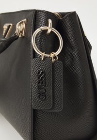Sac à main noir texturé avec matériel doré, comprenant un porte-clés circulaire et une étiquette embossée avec "GUESS". Design structuré.
