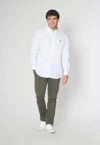 Camicia bianca in cotone con bottoni, maniche lunghe e un piccolo logo sul petto, abbinata a pantaloni verde oliva e sneaker bianche.