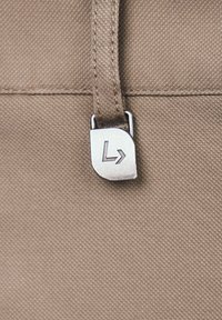 Szczegół beżowej torby z tkaniny z metalowym suwakiem w kolorze srebrnym, który posiada geometryczne logo. Widać teksturowaną powierzchnię.