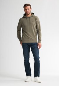 Sudadera verde con capucha, de manga larga y cordones, combinada con jeans oscuros y zapatillas blancas, sobre un fondo claro y uniforme.