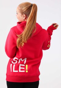 Felpa rossa con tasca frontale, caratterizzata dalla parola "SORRIDI!" in bianco e giallo brillante sul retro. Tessuto morbido e vestibilità comoda.