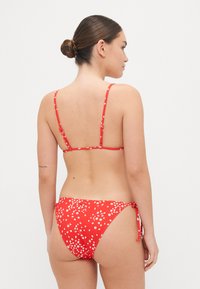 Bikini floral rouge avec de fines bretelles, côtés noués et finition sans couture. Présente de petits motifs de fleurs blanches sur le tissu.