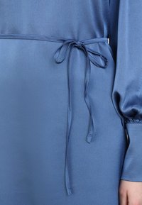 Blauwe jurk met een gladde textuur, voorzien van een stoffen strik in de taille en loszittende mouwen. Het materiaal heeft een subtiele glans.