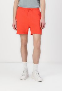 Pantaloni corti in cotone rosso brillante con vita elastica e cordoncino, caratterizzati da una vestibilità comoda e una texture liscia, abbinati a scarpe da ginnastica bianche.