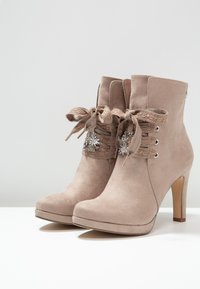 Beige suede high-heeled ankle boots with lace-up ribbon laces and silver flower-shaped charms on a white surface and plain background.