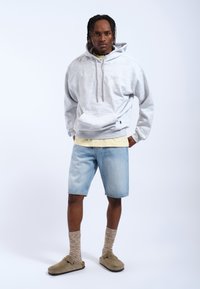 Grijze fleece hoodie met een voorzak, gedragen over een lichtgele shirt. Gekocht met lichtblauwe spijkerbermuda's, bruine slippers en gemusterde sokken.