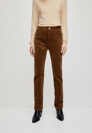 Person iført brune kordbuks, beige langærmet skjorte og sorte ankelstøvler med hæl, stående mod en ensfarvet lys baggrund.