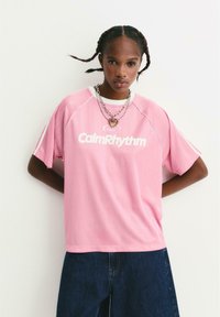 Ung person med fletninger bærer et lagdelt hjertehalskæde, en lyserød "CalmRhythm" t-shirt og mørkeblå jeans, stående mod en ensfarvet væg.