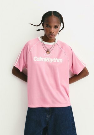 Un jeune avec des cheveux tressés porte un collier en couches en forme de cœur, un t-shirt rose "CalmRhythm" et un jean bleu marine, se tenant devant un mur uni.