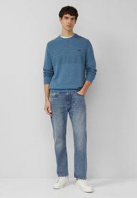 Pull tricoté bleu clair à rayures horizontales texturées, associé à un jean bleu délavé et des baskets blanches, modèle debout devant un fond uni.