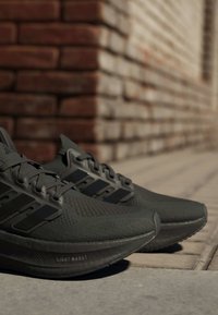 Двойка тъмно сиви маратонки Adidas с подметки "Light Boost", поставени на тротоар до тухлена стена на слънце.