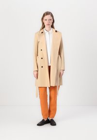 Manteau trench beige avec des boutons noirs, manches longues, ceinture à nouer, porté par-dessus une chemise blanche à col et un pantalon orange, assorti à des chaussures noires.
