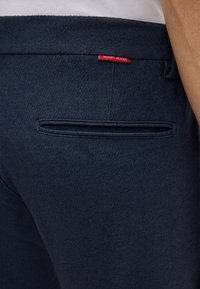 Granatowe spodnie o teksturowanym wykończeniu, z tylną kieszenią i czerwoną metką "JOOP JEANS" podkreślającą paski.