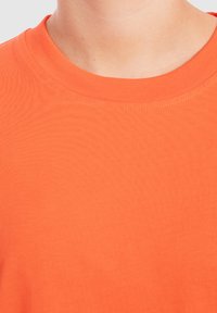 Oranžové triko s kulatým výstřihem vyrobené z žebrované bavlněné tkaniny, které se vyznačuje jemnou texturou a decentním šitím kolem výstřihu.