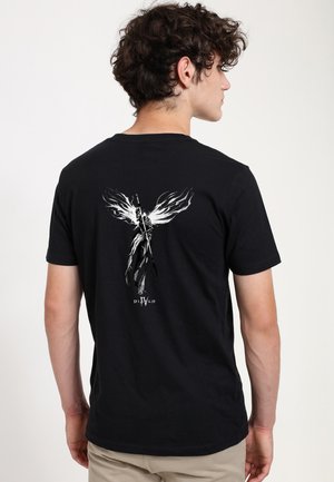 Ung mand i beige bukser iført en sort t-shirt med en hvid ildfyldt figur med vinger, der holder et spyd og teksten "Diablo IV" på ryggen.