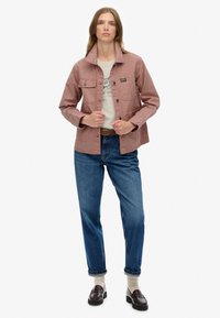 Różowa kanwasowa kurtka z kołnierzem klasycznym, zapinana na guziki, z dwoma kieszeniami na piersiach i swobodnym fasonem, zestawiona z niebieskimi dżinsami i ciemnymi butami.