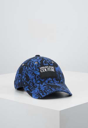 Sort og blåt mønstret baseballkasket med "Versace Jeans Couture" mærkat foran, vist på en hvid overflade mod en grå baggrund.