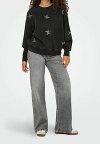 Sweat-shirt noir avec des motifs d'étoiles argentées, associé à un jean à jambes larges gris clair. Le mannequin est debout de manière décontractée, les chaussures visibles en bas.