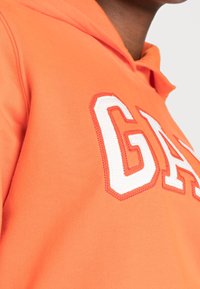 Sudadera naranja de tela suave con un prominente logo bordado en blanco "GAS" con un contorno rojo.