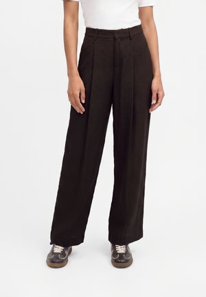Personne portant un pantalon noir à jambes larges, un haut blanc à manches courtes et des baskets marron foncé avec des lacets blancs, debout devant un fond uni.