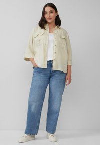 Beige linnen overhemd met twee borstzakken, met knopen; gedragen over een witte top en gecombineerd met lichtblauwe denim jeans en witte schoenen.