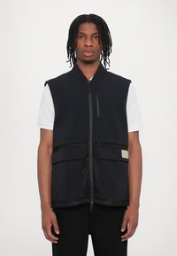 Black sleeveless vest with a zippered front, featuring mesh accents and two front pockets. Made from lightweight fabric, textured finish.