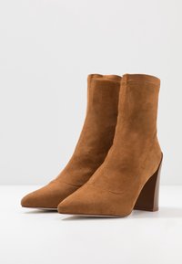 Bottines en suede marron avec des pointes aiguës et des talons hauts épais, posées côte à côte sur une surface blanche.