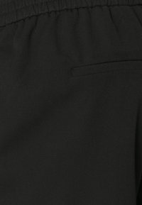 Tissu noir avec une surface légèrement texturée. Dispose d'une taille élastique et d'une petite poche horizontale sur le côté.