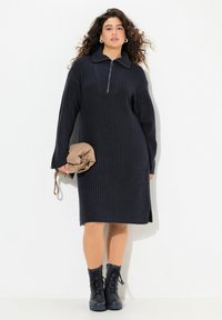 Robe tricotée en marine foncé avec col et zip quart, texture côtelée, fente latérale, associée à une pochette beige et des bottines noires à lacets.