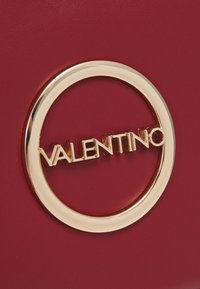 Borsa a mano in pelle rossa con un grande logo circolare dorato con la scritta "Valentino". Texture liscia con accenti lucidi.
