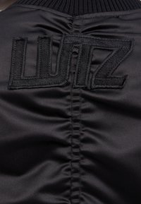 Schwarze Satin-Bomberjacke mit gerafften Nähten und auffälligem gesticktem "LUTZ"-Logo auf dem Rücken. Textilstruktur verleiht Tiefe.