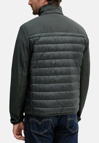 Giacca imbottita verde scuro, caratterizzata da una texture trapuntata sul retro e da un tessuto liscio sulla parte anteriore. Colletto alto e design aderente.