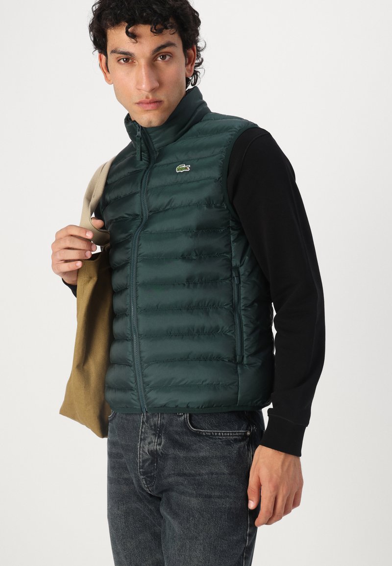 Zielona kamizelka z ociepleniem, z poziomymi przeszyciami, zamek błyskawiczny i osłona na brodę. Logo Lacoste na lewej piersi. Model trzyma brązowy ciuch.