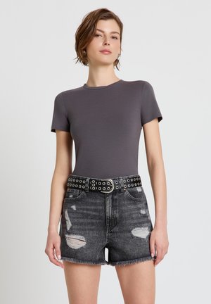 Donna con capelli castani corti che indossa una maglietta grigia aderente, pantaloncini in denim neri consumati e una cintura nera con occhielli, in piedi davanti a uno sfondo bianco.