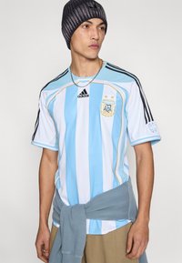 Ποδόσφαιρο της Αργεντινής με μπλε και λευκές κάθετες ρίγες, με κεντημένο λογότυπο, μαύρες λεπτομέρειες και άνετη εφαρμογή. Γκρι πουλόβερ δεμένο γύρω από τη μέση.