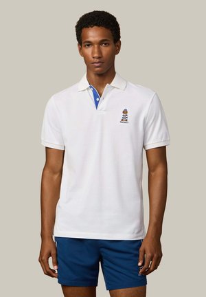 Jonge man staand, gekleed in een wit poloshirt met een blauwe kraagdetail en een geborduurde beer, gecombineerd met marineblauwe korte broek.