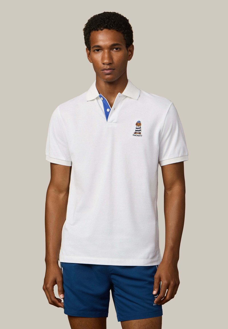 Joven de pie que lleva una camisa polo blanca con detalle en el cuello azul y un oso bordado, combinado con pantalones cortos azul marino.