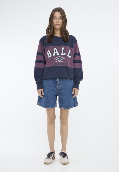 Pulover de tip crewneck în navy și burgundy cu textul "BALL 1974"; asortat cu șorturi din denim și adidași bej. Dispune de mâneci în dungi.