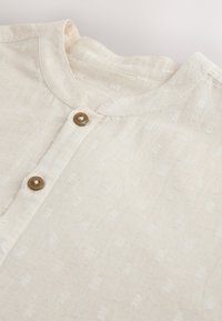 Light beige shirt with round neckline and two brown buttons on the placket, showing subtle textured fabric pattern.