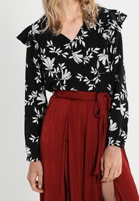 Blusa nera con motivo floreale bianco, scollatura a V e spalle arricciate, abbinata a una gonna rossa fluida a vita alta con dettaglio di lacci.