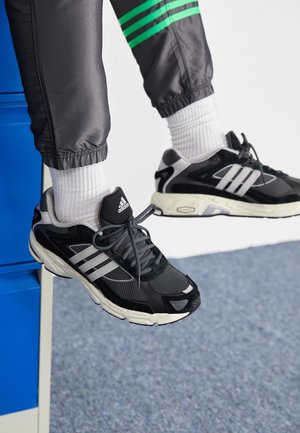 Pieds portant des baskets Adidas noires et blanches et des chaussettes blanches, jambes en pantalon noir avec des bandes vertes, flottant au-dessus d'un tapis bleu et d'une armoire bleue.
