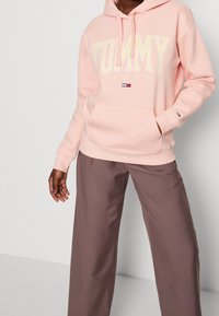 Sudadera rosa con "TOMMY" y "JEANS" impresos en amarillo, con un bolsillo frontal y cordones ajustables. Se lleva con pantalones marrón claro.