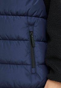Marineblå puffervest med sidelommer med sort hardware, quiltet tekstur og kontrasterende sorte fleeceærmematerialer.