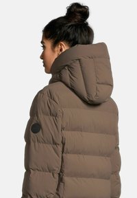 Braune Pufferjacke mit einer hohen, gepolsterten Kapuze und gesteppter Optik. Sie verfügt über ein rundes Logo-Patch am linken Ärmel. Weiche Textur und warmes Erscheinungsbild.