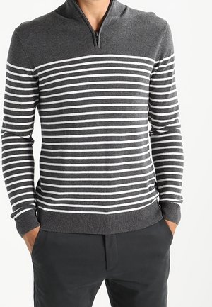 Homme portant un pull à rayures horizontales gris foncé et blanc avec un col à fermeture éclair courte et un pantalon noir, les mains dans les poches.